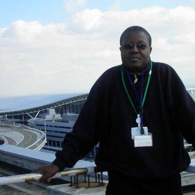 photograph of Calistus Wachana standing in front of a large building perhaps a stadium or airport. A young black man with short hair and wearing glasses