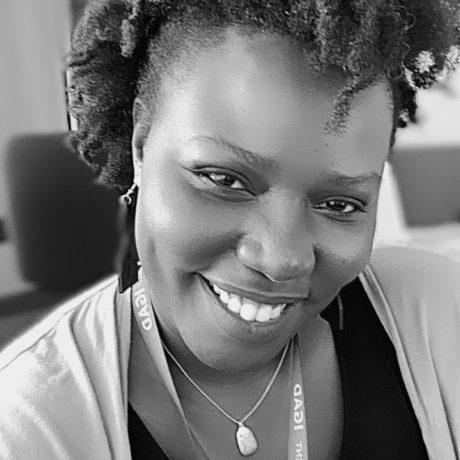 a black and white head and shoulders photo of Linda Ogallo a young black woman with mid length hair, wearing a dark top and lighter cardigan and an ICPAC lanyard