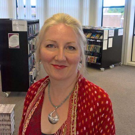 A white woman with blond hair tied back, wearing a red jacket and necklace