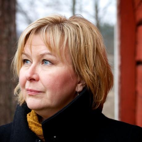 A white woman with short blond hair wearing a dark coat and yellow scarf