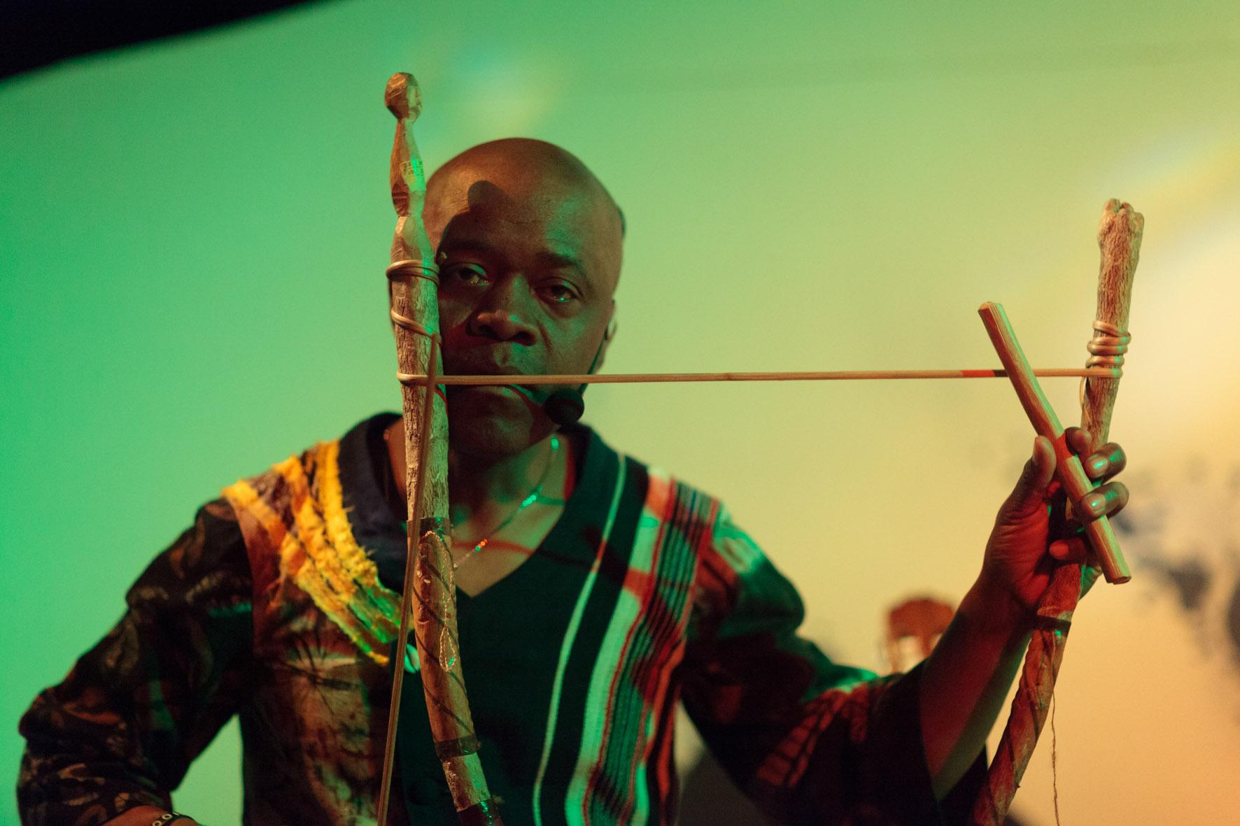 a black man wearing a black and gold tunic and playing the dodo mouth bow