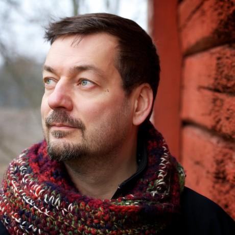 A white man with short dark hair and a beard, wearing a multi coloured scarf