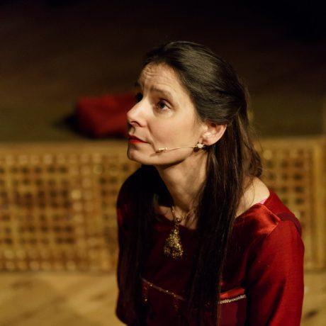 Xanthe in costume for The Shahnameh, long dark hair and red dress sitting on stage looking to one side