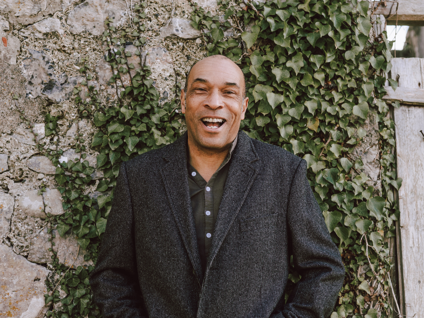 Phil Okwedy is standing in front a wall wearing a dark grey jacket and green top. The wall has some climbing ivy in the background and an old door to the right.