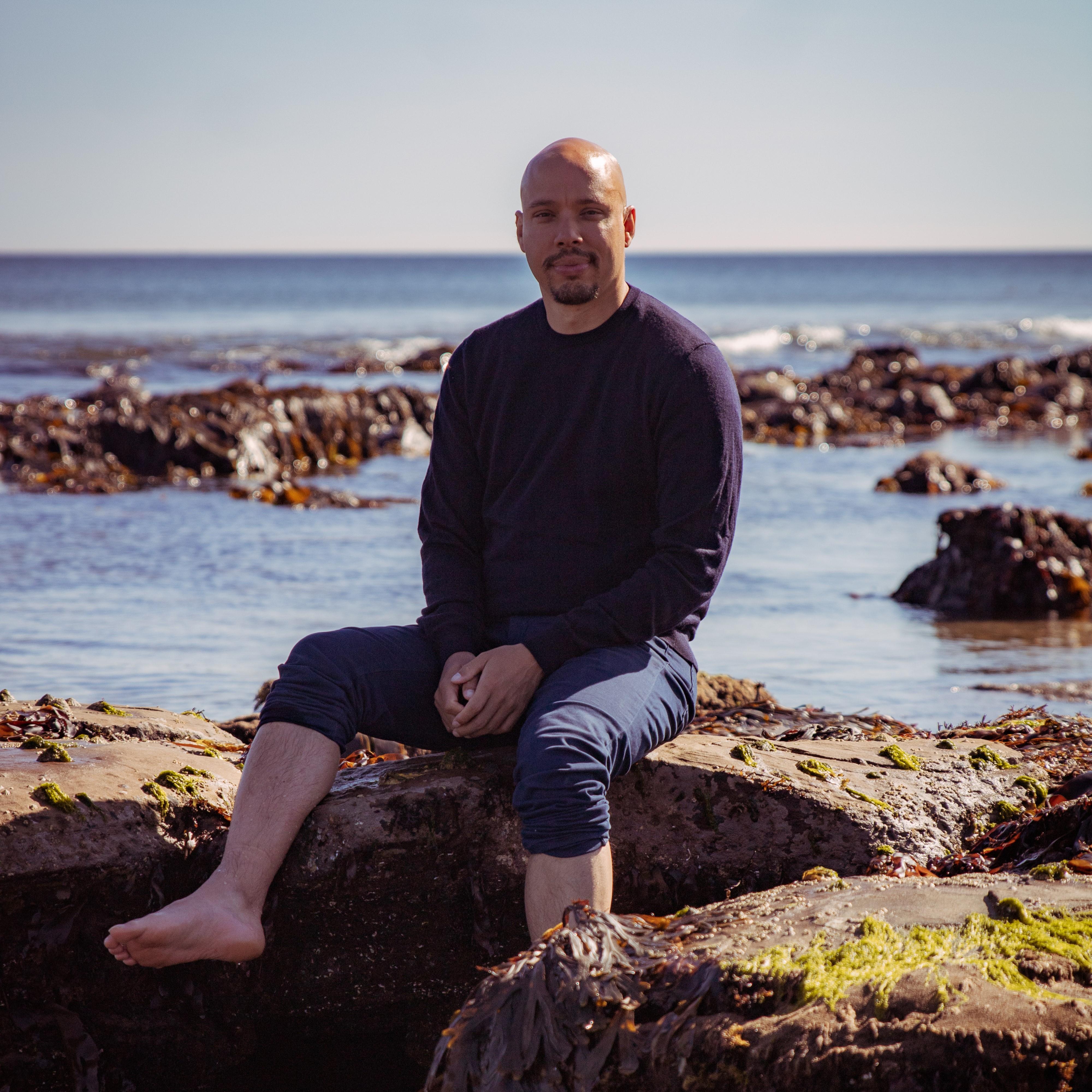 Chris is sat on rocks near the sea.