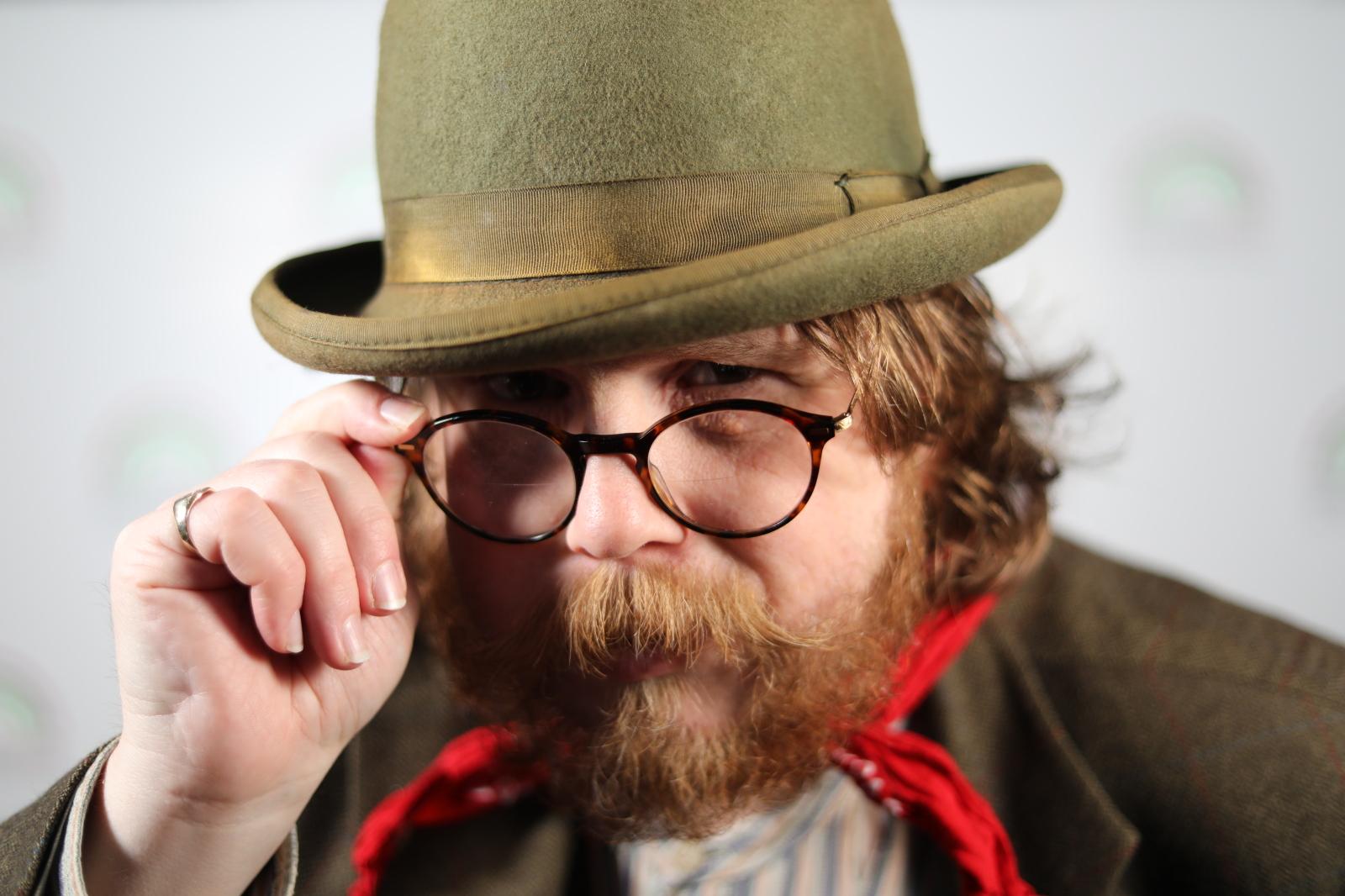 Chris is wearing a dark green bowler hat, glasses and holding his hand up to his face