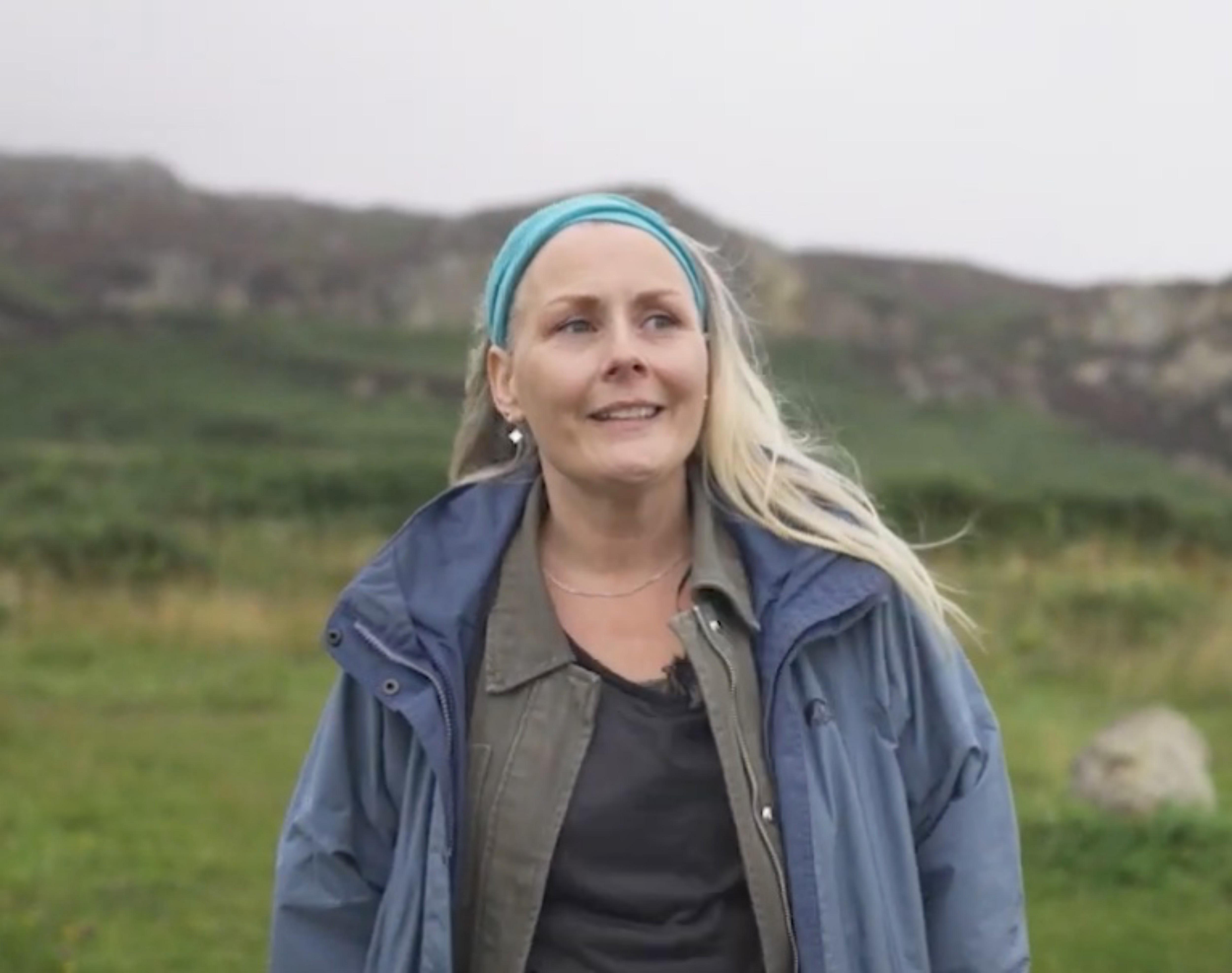 Gillian has long blonde hair flowing in the wind. She is wearing a blue coat against the backdrop of green rocky hills.