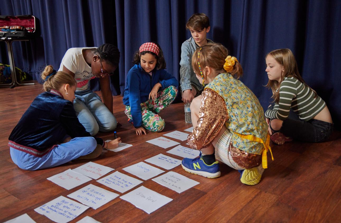 The young participants are sat on a wooden floor with pieces of paper and learning how to create a narrative of a story.