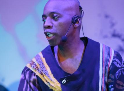 a black man wearing a black and gold tunic and playing a drum