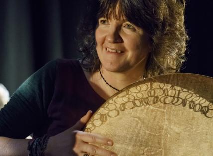 a white woman with shoulder length brown hair, playing a large drum