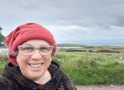 Photo of Mair standing in front of the North Wales moors, wearing a red hat and glasses