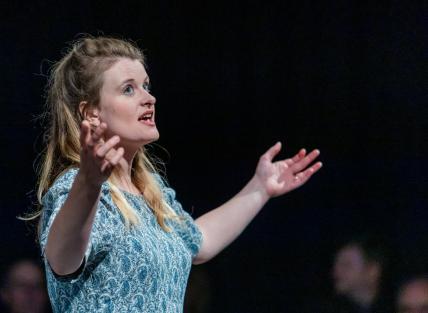Photo of Tamar in a blue dress, long blonde hair, arms reaching outwards to the audience. 