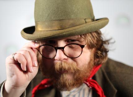 Chris is wearing a dark green bowler hat, glasses and holding his hand up to his face