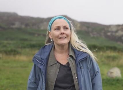 Gillian has long blonde hair flowing in the wind. She is wearing a blue coat against the backdrop of green rocky hills.