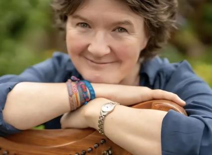 Stacey is leaning on a wooden harp and has short brown hair.